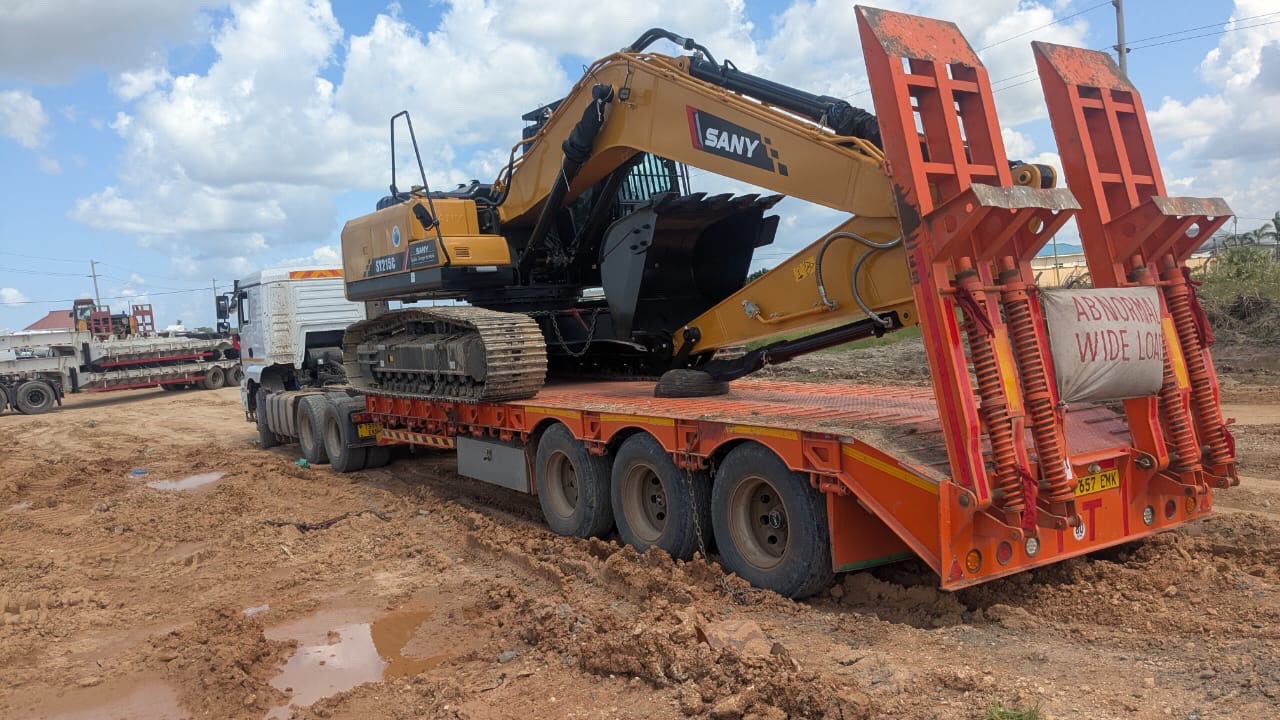 A yellow SANY SY215G crawler excavator chained to an orange lowbed trailer marked abnormal wide load, on a muddy African road